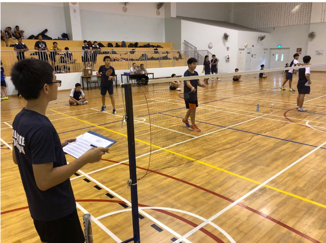 Students playing badminton in an indoor gymnasium with a net, spectators in bleachers, and a referee holding a clipboard.